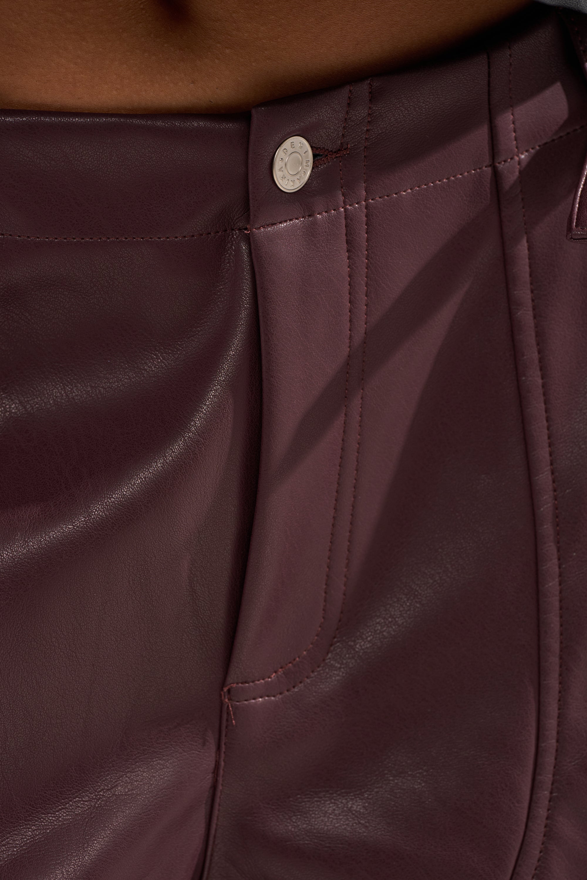 Close-up of a person wearing the OVER DONE FAUX LEATHER BERMUDA SHORT, featuring a high-waisted fit in dark maroon faux leather with a silver button, detailed stitching at waistband and pocket, and a smooth, shiny finish for a trendy look.