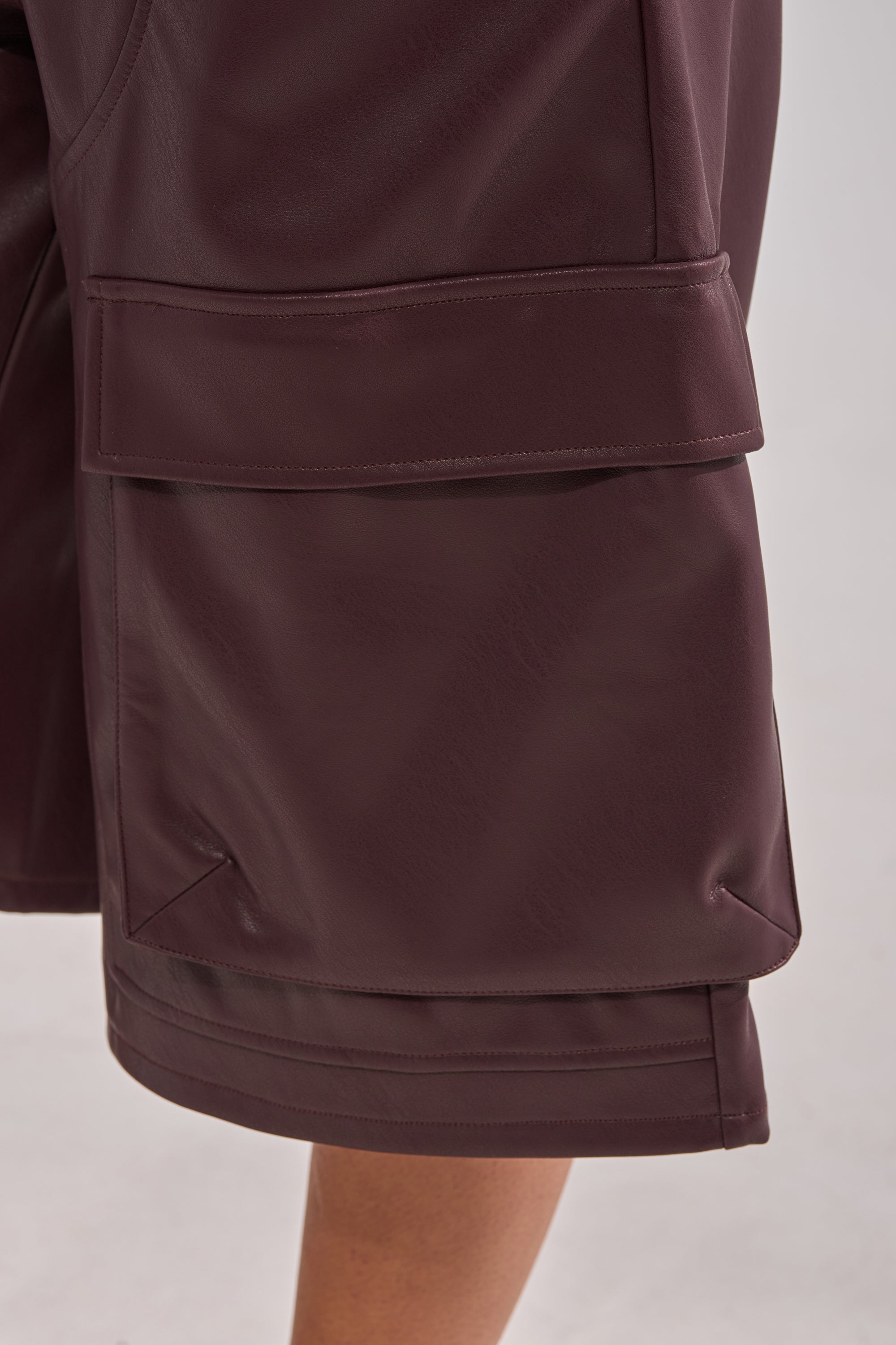 A close-up shows a person wearing the OVER DONE FAUX LEATHER BERMUDA SHORT in dark brown, featuring large cargo pockets and a smooth faux leather texture, with part of their leg visible against a plain background.