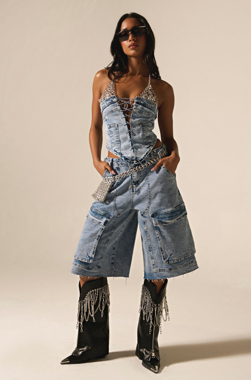 Wearing acid wash denim corset and LANA DENIM JORT with cargo pockets, black cowboy boots with chains, long dark hair, sunglasses, hands in pockets against a plain background.