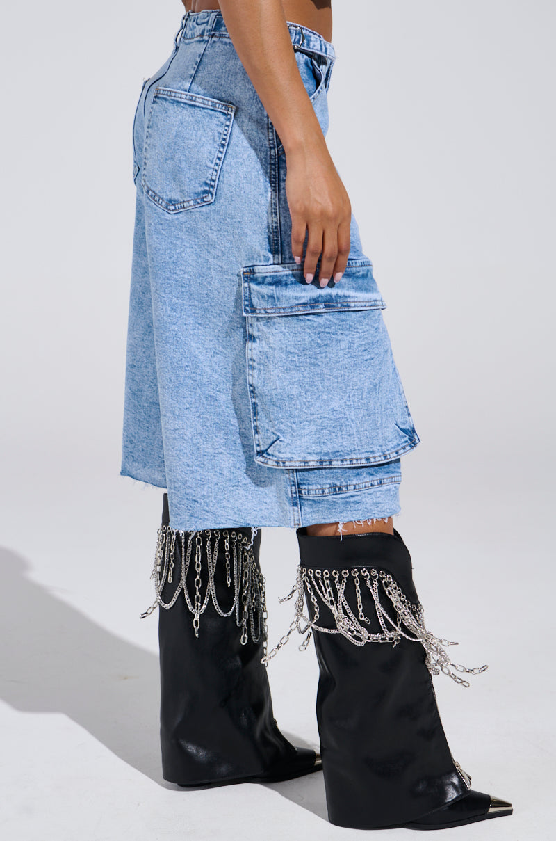 Someone is wearing the LANA DENIM JORT in light blue acid wash, featuring large side pockets. They're paired with black high-heeled boots adorned with silver chains draping down. The background is plain and minimalistic.