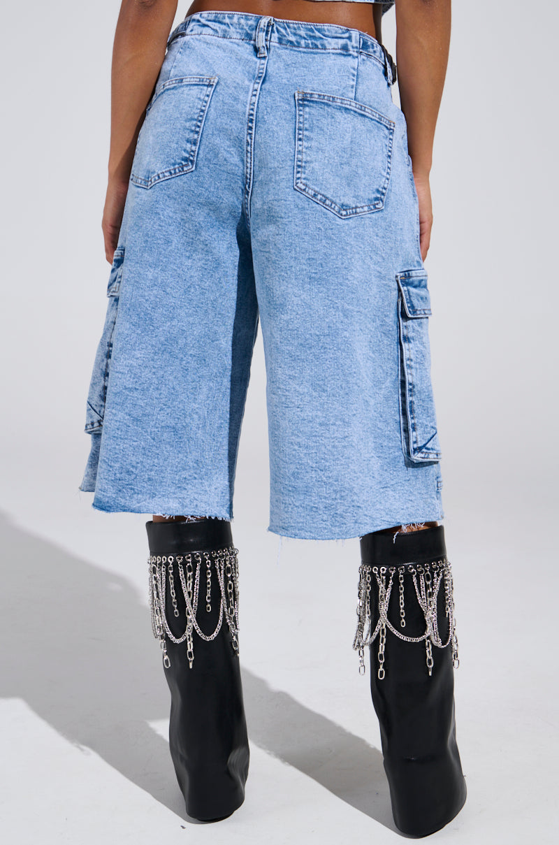 A person in the light blue acid wash LANA DENIM JORT with frayed hems faces away from the camera, pairing these stylish shorts with black high-heeled boots adorned with silver chains.