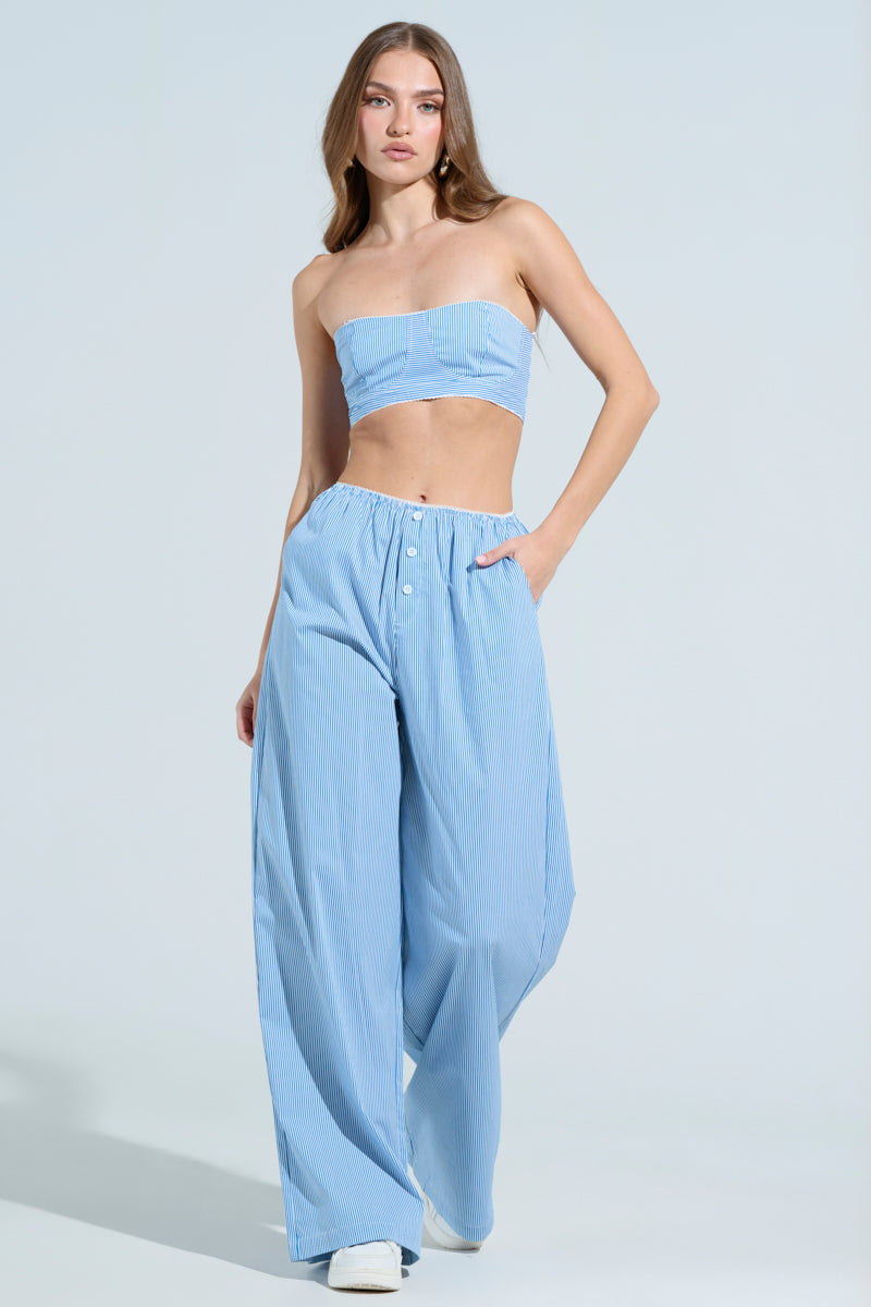 A person in a light blue, strapless crop top and matching high-rise DAY AND NIGHT PANTS with a subtle pinstripe design poses gracefully, long hair flowing against a light gray background.