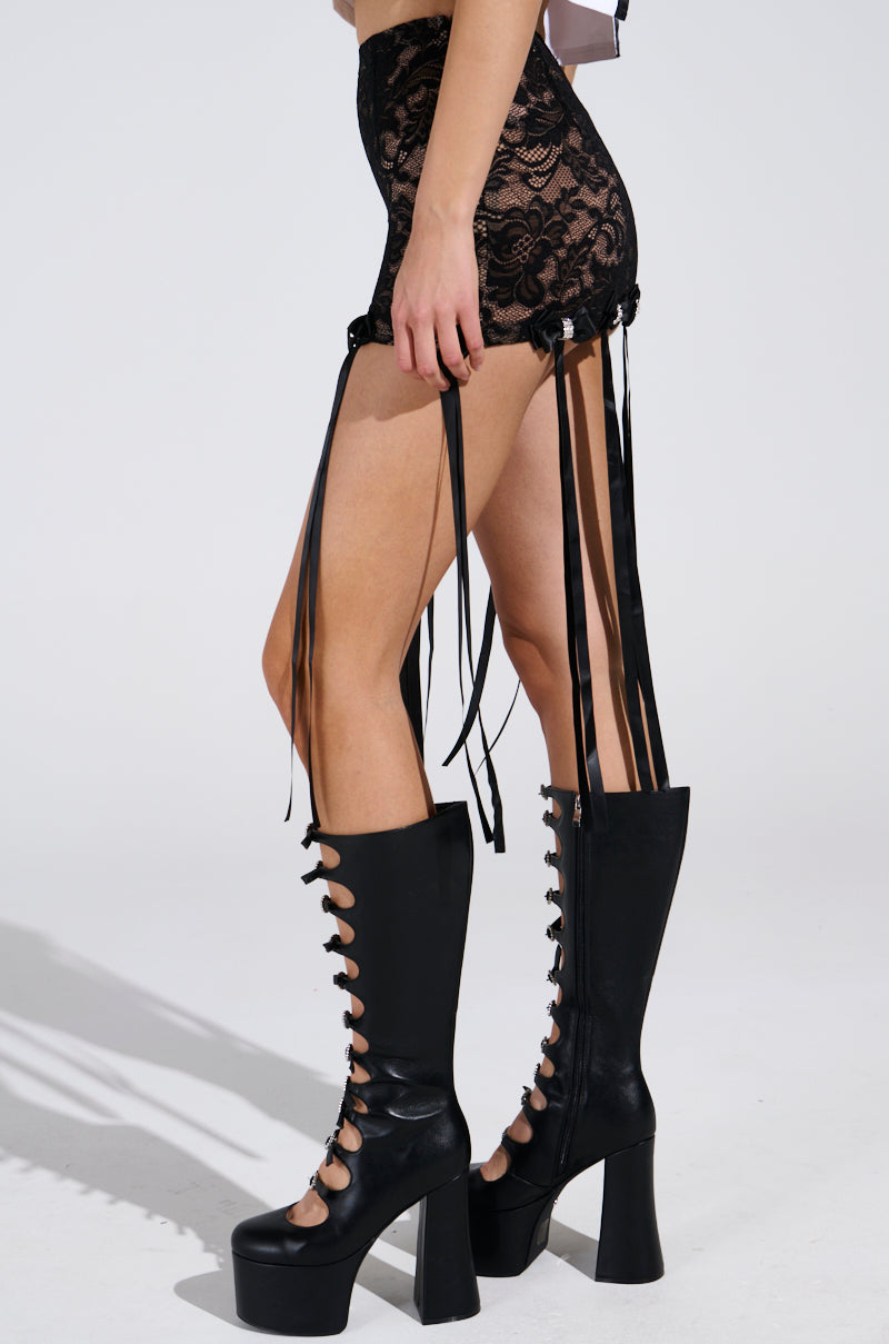 A person wearing the GIRLY POP LACE MINI SKIRT, enhanced with satin bow appliques, is paired with tall black platform boots featuring lace-up fronts and chunky heels. They stand against a plain, light background that highlights the outfit.