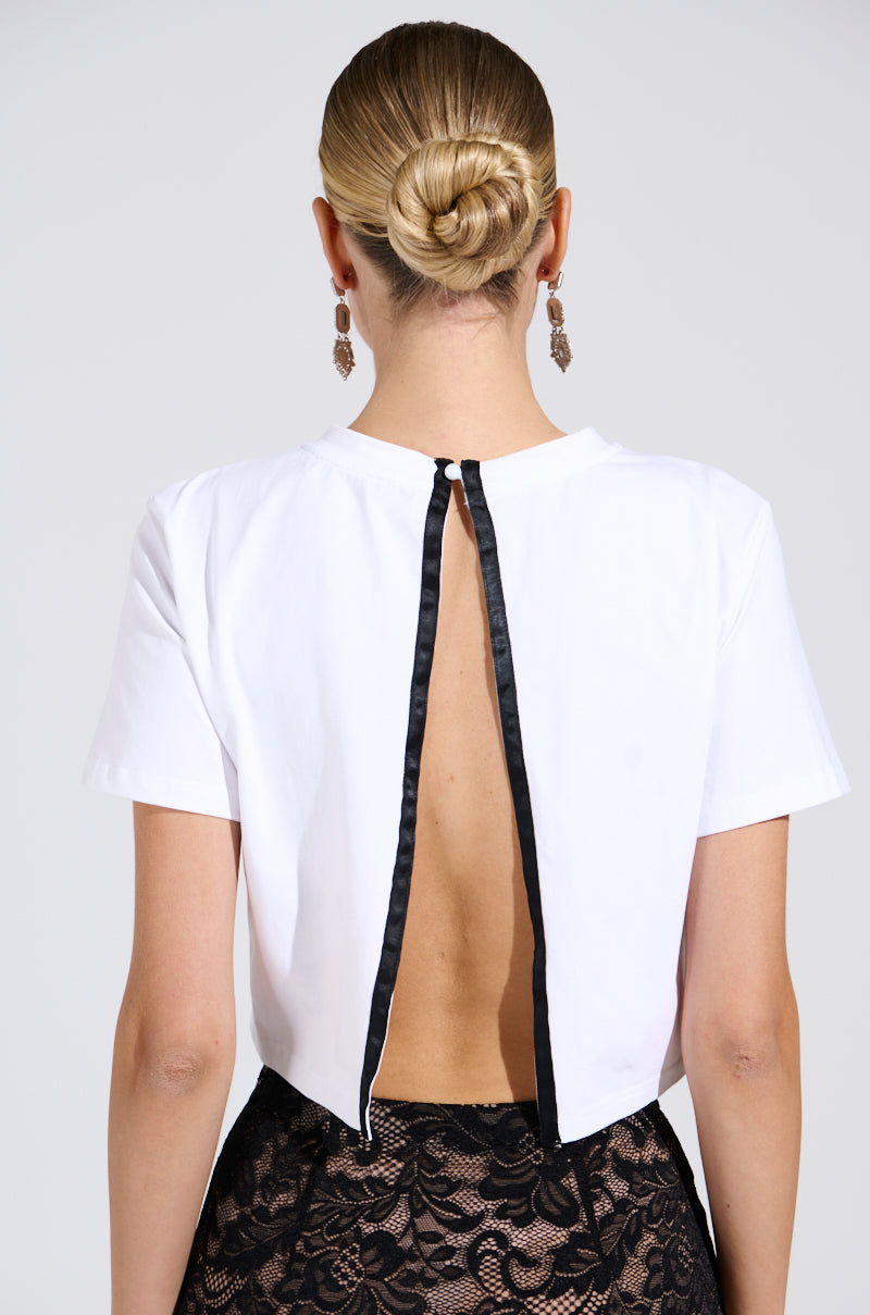 A person with a neat bun is shown from behind in the GIRLY POP BOW TEE, a white open-back top secured by a lacy bow graphic, paired with a black lace skirt and dangling earrings against a plain white background.