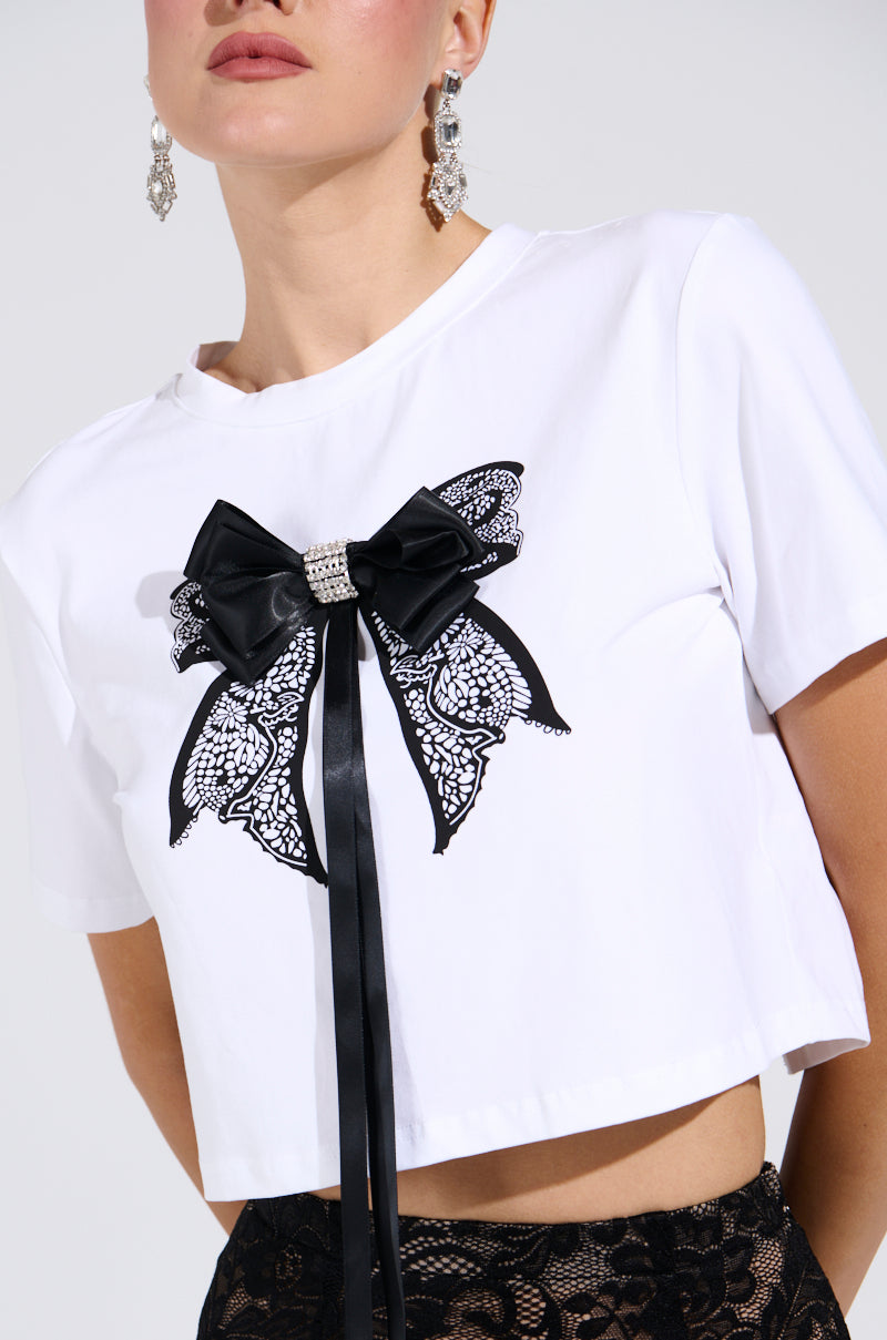 Wearing a GIRLY POP BOW TEE with its black lace and satin-trimmed open-back design, someone accessorizes with silver dangling earrings against a plain white background, and pairs it with black lace below.