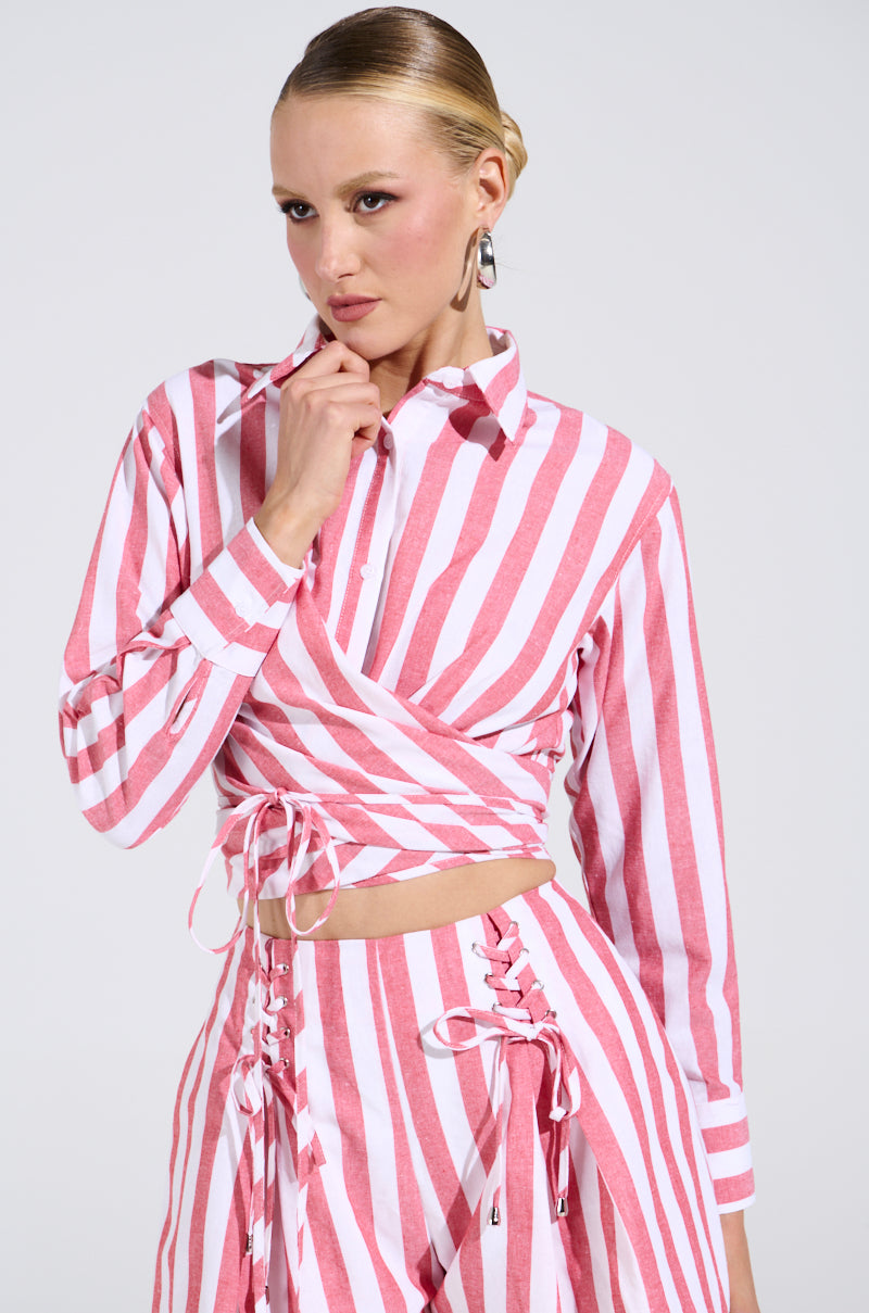 The individual is wearing the GIRL BYE TIE FRONT STRIPE BLOUSE with lace-up pants, featuring a beachy stripe pattern. Posing against a plain background, one hand gently touches their chin.