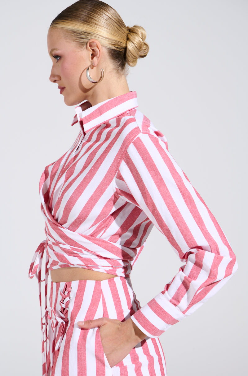 A person with blonde hair in a bun wears the GIRL BYE TIE FRONT STRIPE BLOUSE in pink and white, paired with matching pants. They look to the side against a light gray background, exuding casual elegance and a touch of linen style charm.