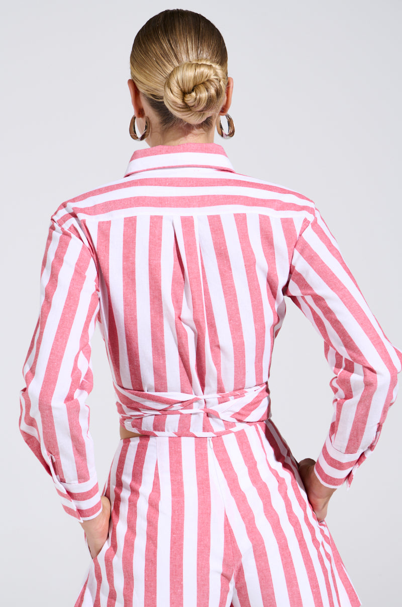 From behind, a person with blond hair in a bun wears the GIRL BYE TIE FRONT STRIPE BLOUSE with pink and white beachy stripes. The ensemble includes a collared top, long sleeves, and matching pants, all paired with large hoop earrings that catch the eye.
