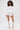 A woman with curly hair wears a white AKIRA Label off-the-shoulder blouse, PLAY WITH ME SHORT high-rise shorts, and white knee-high lace-up heeled boots. She holds a matching clutch and stands against a plain light background.