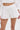 A person stands against a plain background, wearing PLAY WITH ME SHORT high-rise white flowy shorts with a delicate hemline, a white long-sleeve top, and holding a small white purse.