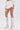 A person wearing a white cropped top, PLAY WITH ME SHORT high-rise flowy shorts, white knee-high lace-up stiletto boots, and holding a small white handbag stands against a plain white background.