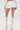 A person stands facing away, dressed in a white cropped top and the PLAY WITH ME SHORT—AKIRA Label’s high-rise shorts with a flowy hemline—paired with knee-high strappy high-heeled boots and a small white bag on a plain white background.