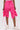 A person models the GIRL ON FIRE RUCHED JOGGER—bright pink, knee-length shorts with adjustable drawstrings—paired with white sneakers against a plain background, highlighting bold streetwear style.