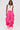 A woman stands confidently against a plain light background wearing the GIRL ON FIRE RUCHED JOGGER in bright pink, a white cut-out tank top, and white sneakers.