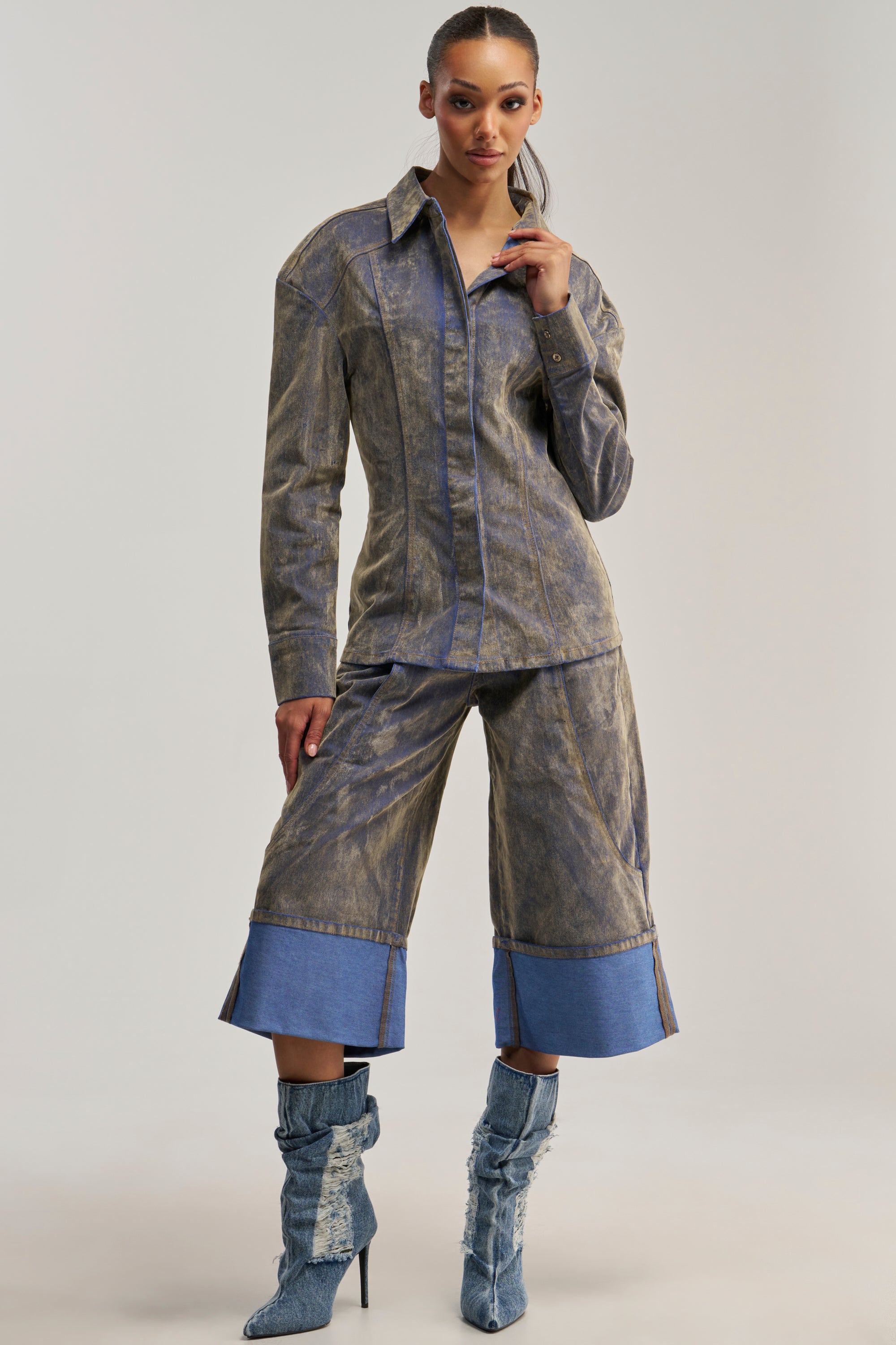 A woman showcases the FALL VIBES VELVET DENIM BERMUDA SHORT IN TAUPE, styled with a button-up shirt and matching high-heeled boots. She stands confidently against a plain background.