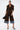 A woman embraces fall fashion in the FALL VIBES VELVET BERMUDA SHORT, styled with a matching brown button-up shirt, denim-cuffed knee-length shorts, a black handbag, pointed heels, silver bracelets, and slicked-back hair.