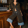 A woman confidently poses by a bar in FALL VIBES VELVET BERMUDA SHORTS, styled with a brown jacket and tan fur-accented heels, embodying chic fall fashion in a dimly lit wooden interior.