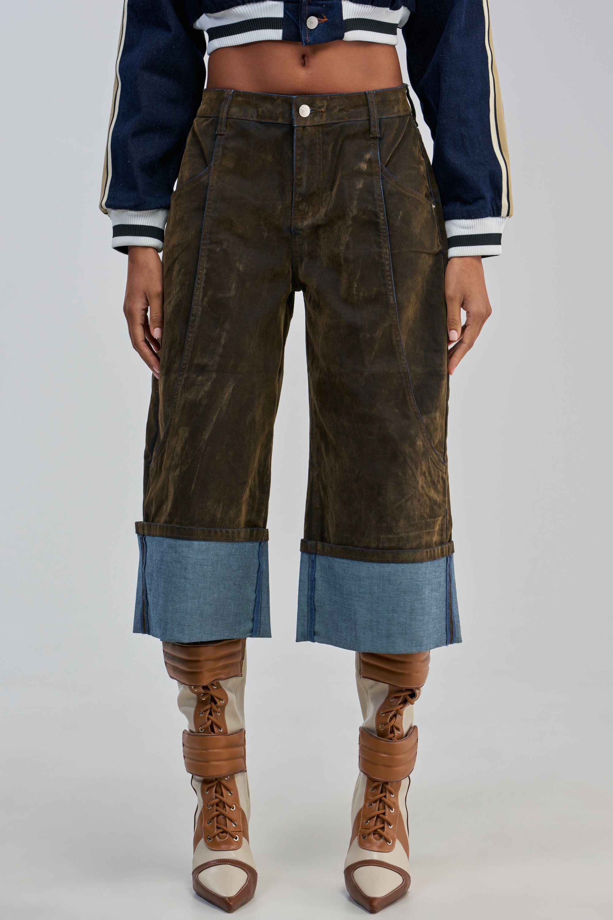 A model wears the FALL VIBES VELVET BERMUDA SHORT—dark brown velvet bermuda shorts featuring wide, light-blue denim cuffs—styled with a cropped, blue-and-white varsity jacket and tall tan-and-cream lace-up boots, set against a plain grey background.