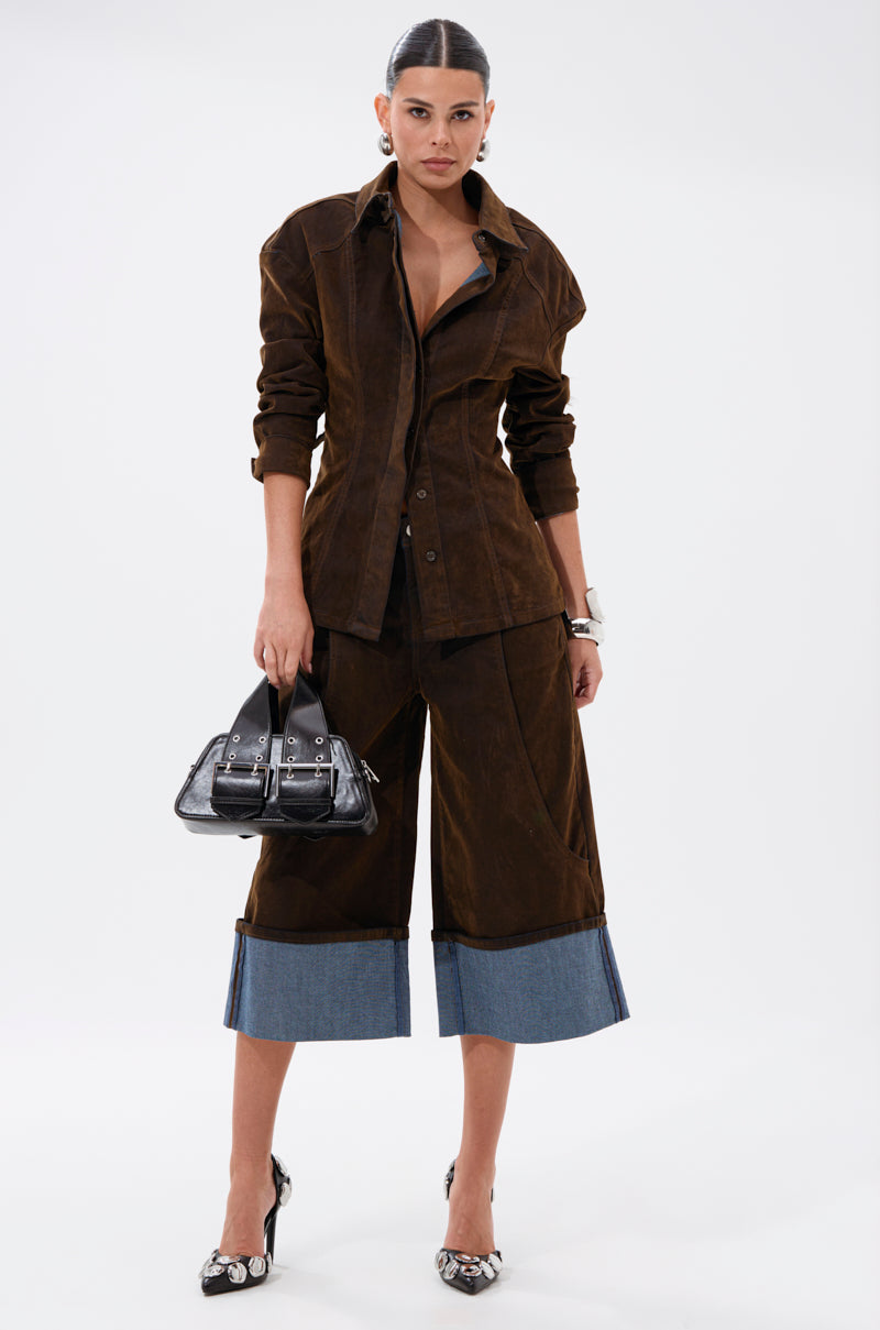 A woman stands against a white background wearing the FALL VIBES VELVET BERMUDA SHORT, styled with a dark brown jacket, blue denim-cuffed details, a black handbag, floral-pointed heels, and slicked-back hair—an effortlessly chic fall look.