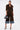 A woman stands against a white background wearing the FALL VIBES VELVET BERMUDA SHORT, styled with a dark brown jacket, blue denim-cuffed details, a black handbag, floral-pointed heels, and slicked-back hair—an effortlessly chic fall look.