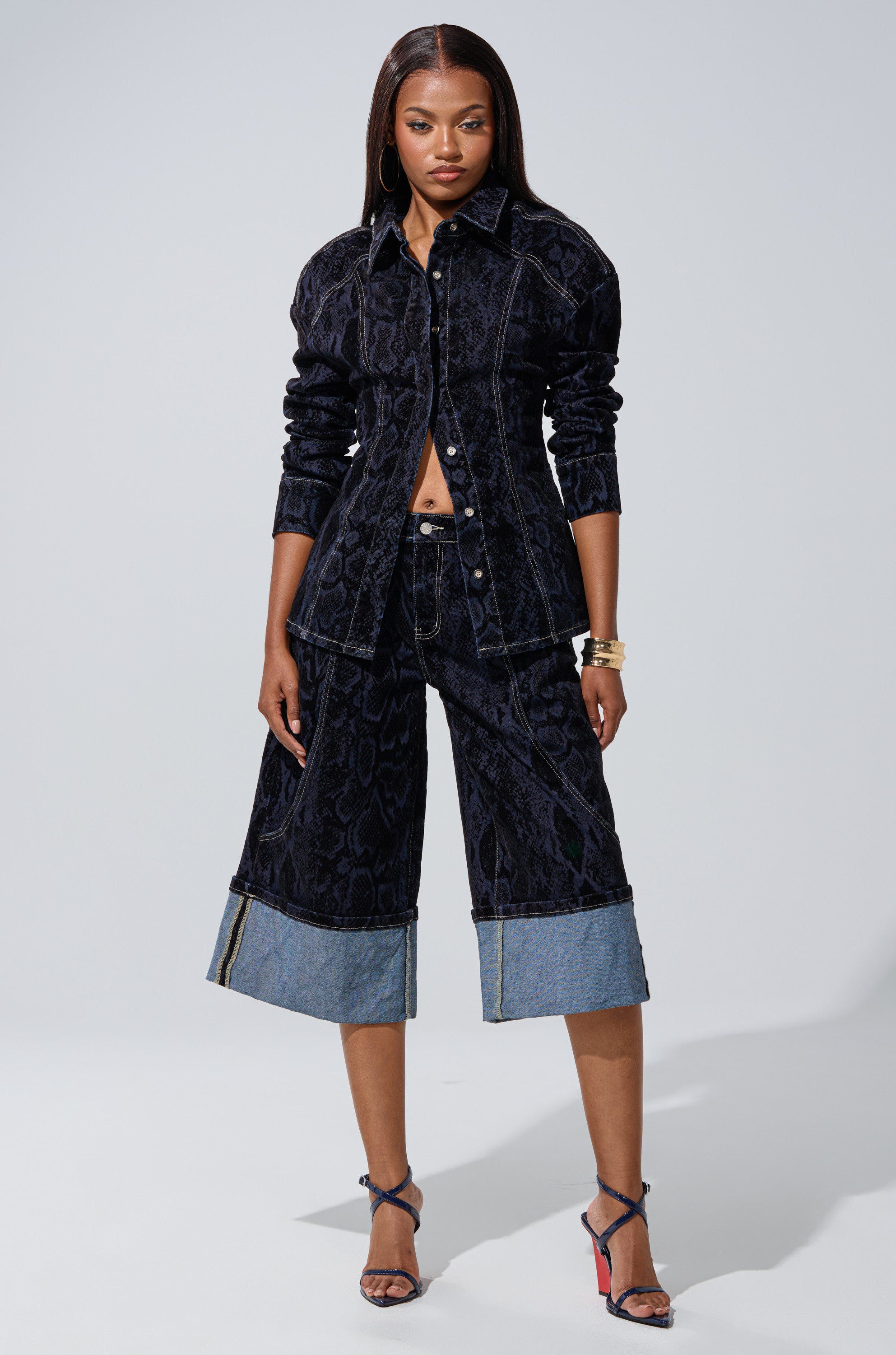 A woman poses confidently in the FEELS LIKE MONEY DENIM BERMUDA SHORT, styled with a dark wash denim shirt, strappy heeled sandals, and gold bangles against a plain background.