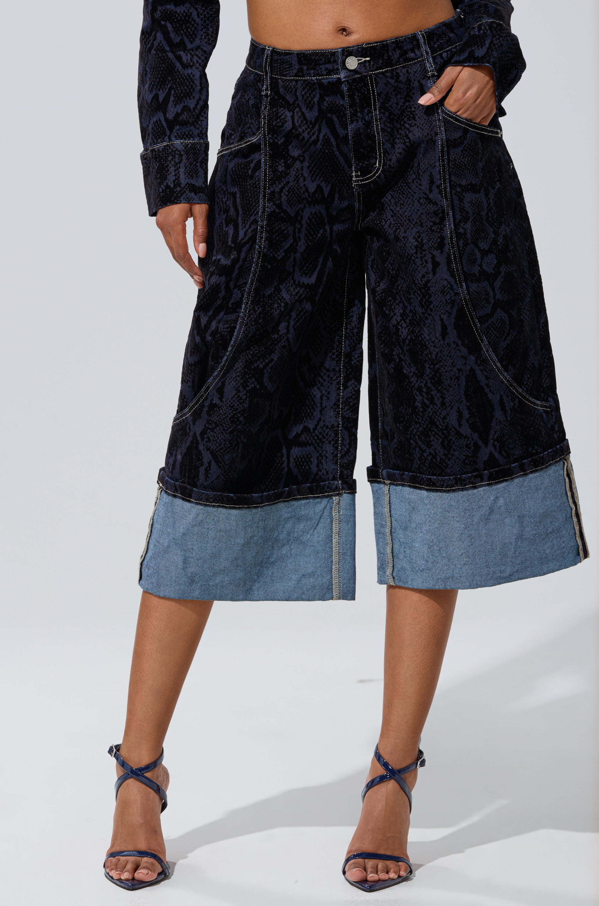 A person wears the FEELS LIKE MONEY DENIM BERMUDA SHORT with strappy dark high-heeled sandals, standing against a plain background.