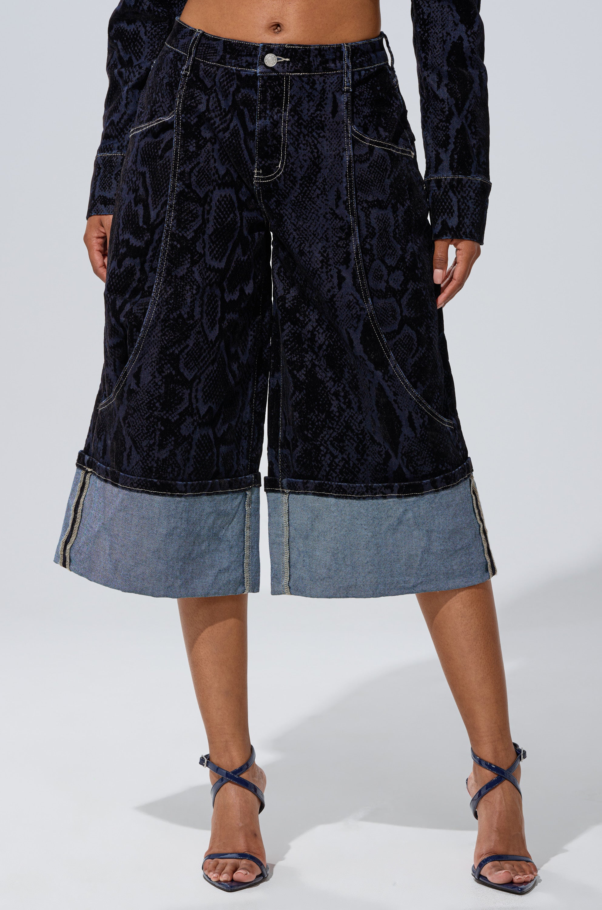 Wearing the FEELS LIKE MONEY DENIM BERMUDA SHORT and blue high-heeled sandals, a person stands against a plain light background.