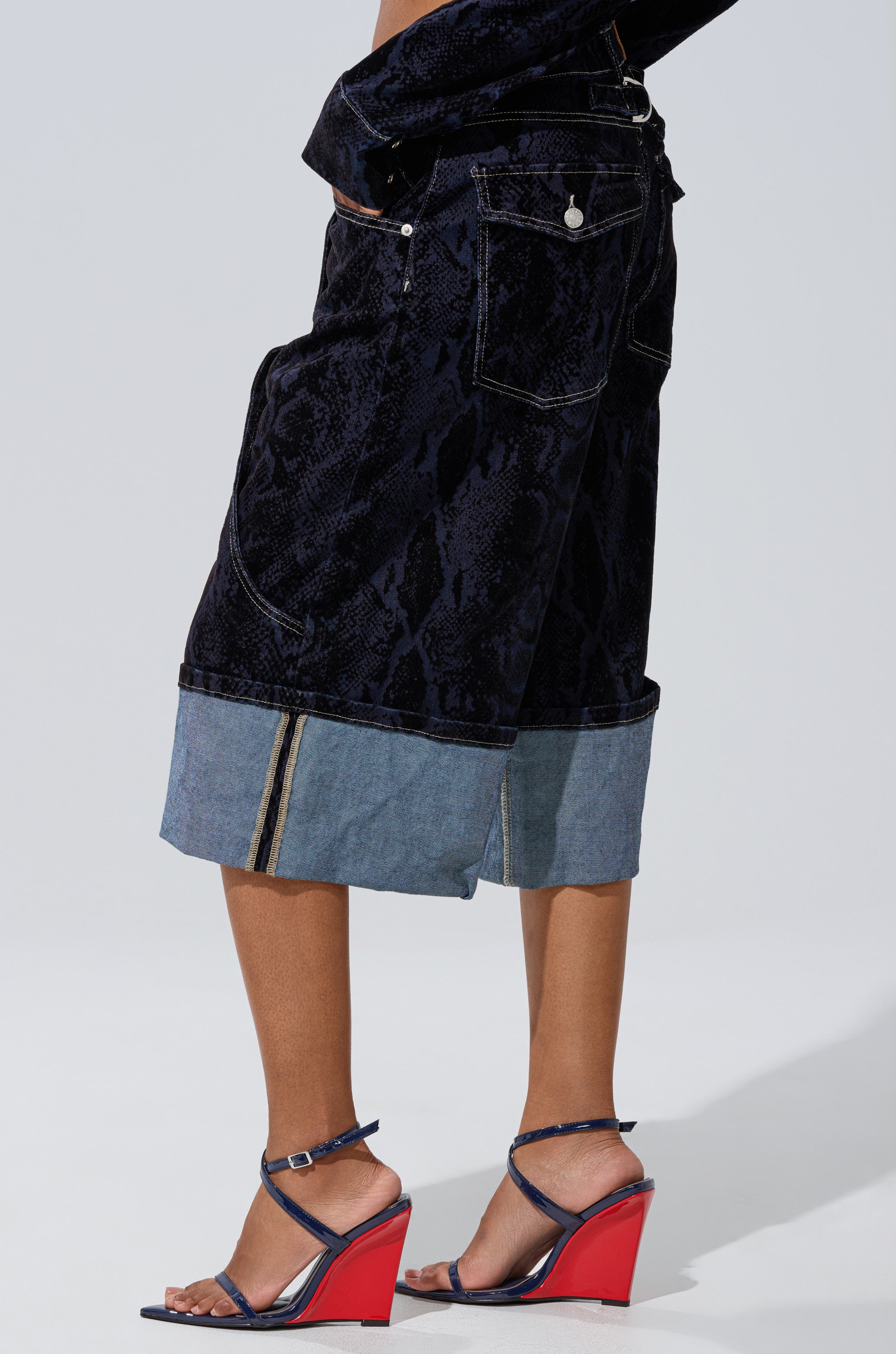 A person wearing the FEELS LIKE MONEY DENIM BERMUDA SHORT, featuring a high waist and dark wash with a light denim cuff, pairs them with navy and red wedge sandals while standing against a plain light background.