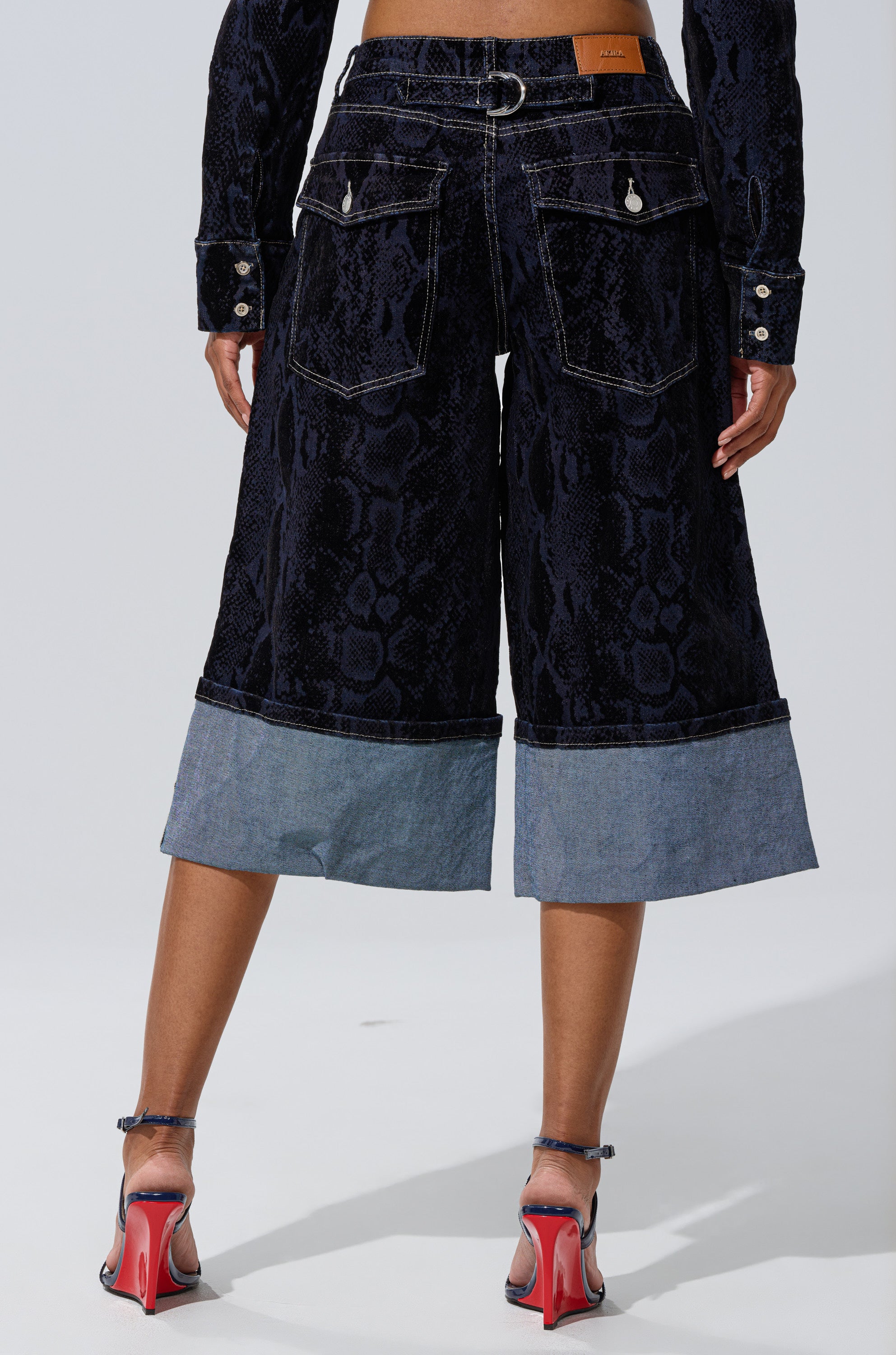 A person stands facing away, wearing the FEELS LIKE MONEY DENIM BERMUDA SHORT—cropped dark wash denim with a snakeskin pattern, large back pockets, cuffed hems—paired with high-heeled open-toe shoes and a matching jacket.