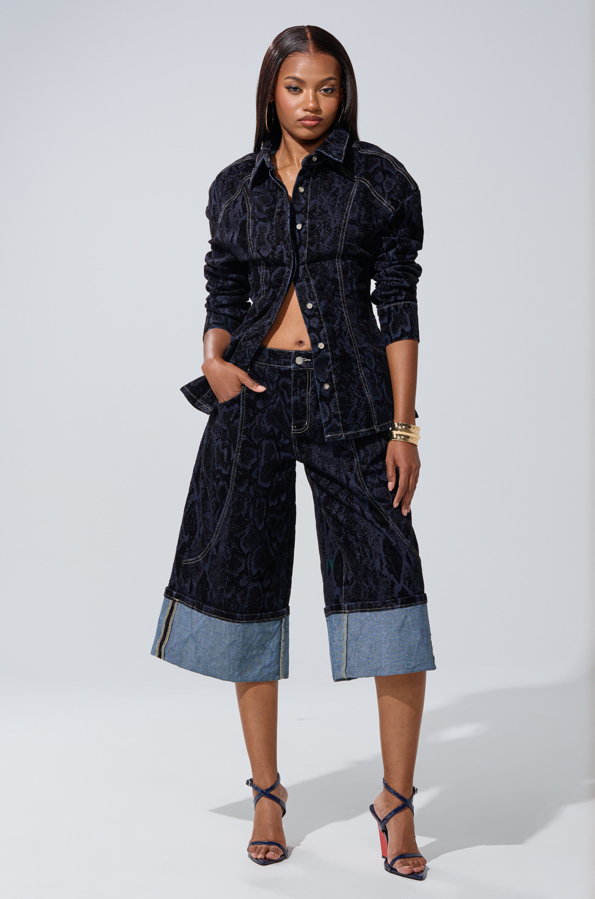 A woman stands confidently wearing the FEELS LIKE MONEY DENIM BERMUDA SHORT in a dark wash, styled with strappy heels and bracelets for a chic, modern look.