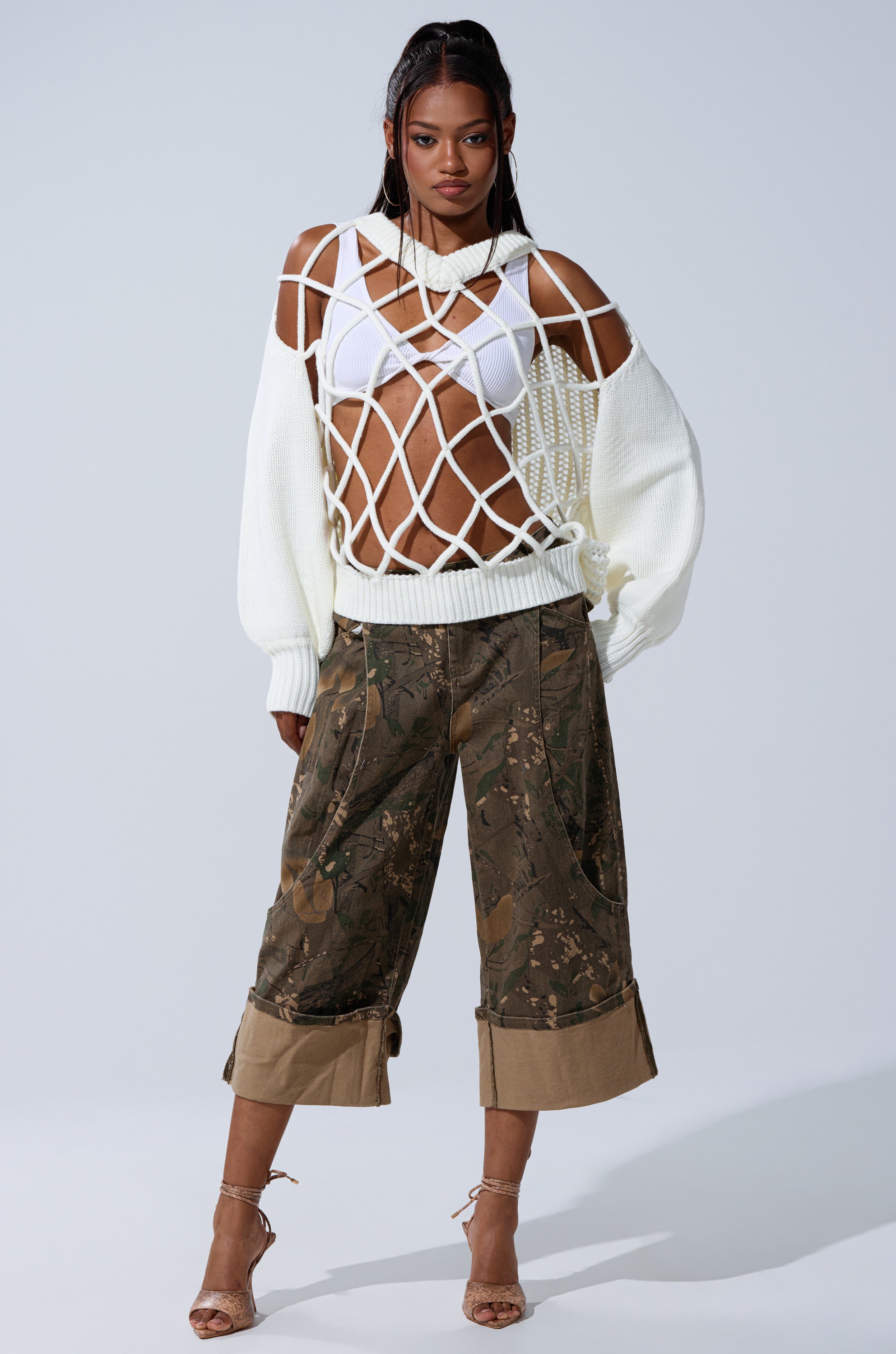 A woman poses in the DOWN AND DIRTY CAMO BERMUDA SHORT paired with a white netted sweater, white bikini top, and strappy heels against a plain light background. 1