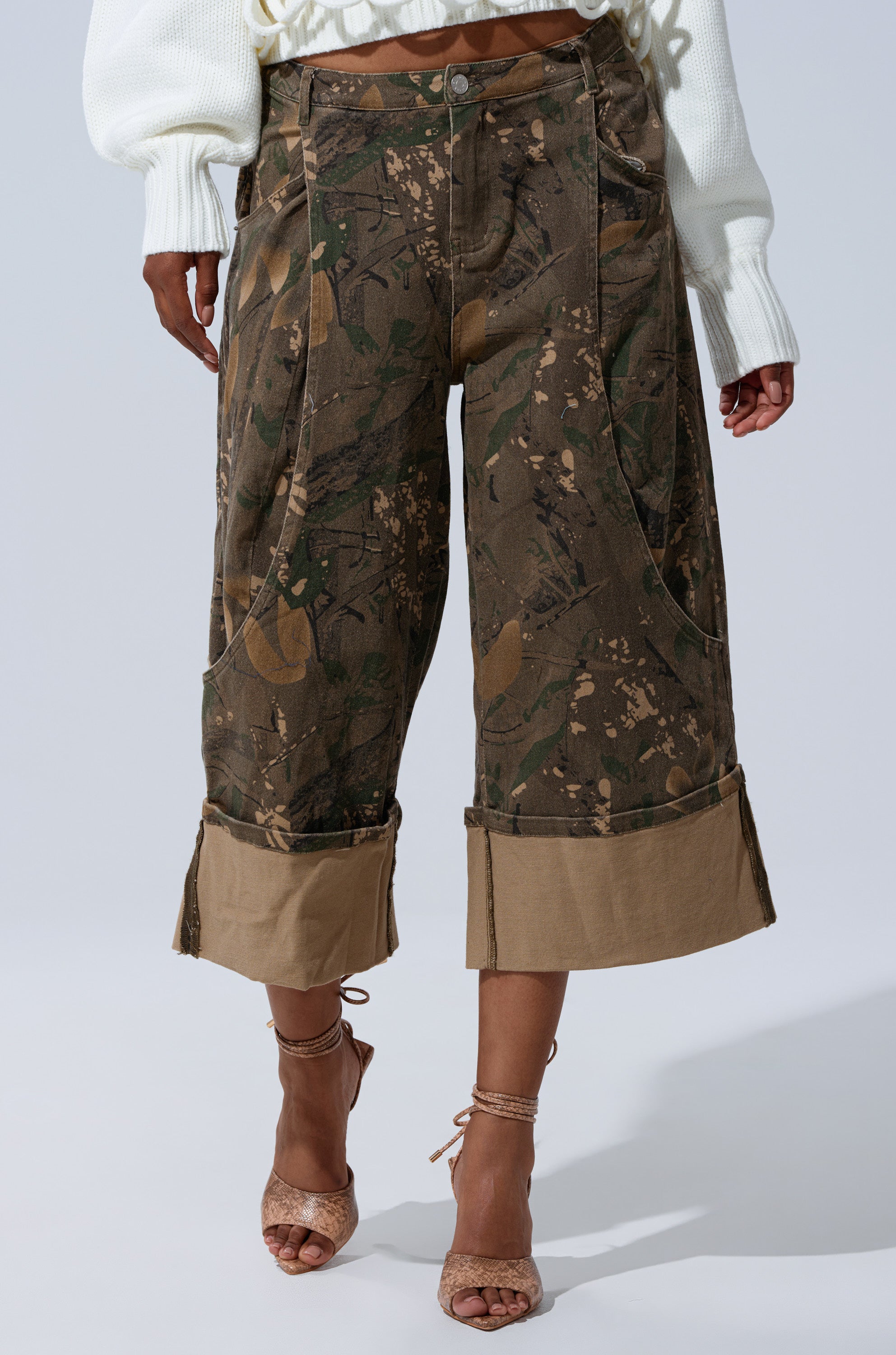 Someone poses confidently against a plain light background, wearing the DOWN AND DIRTY CAMO BERMUDA SHORT with a mid-rise fit, paired with a white sweater and strappy high-heeled sandals. 0