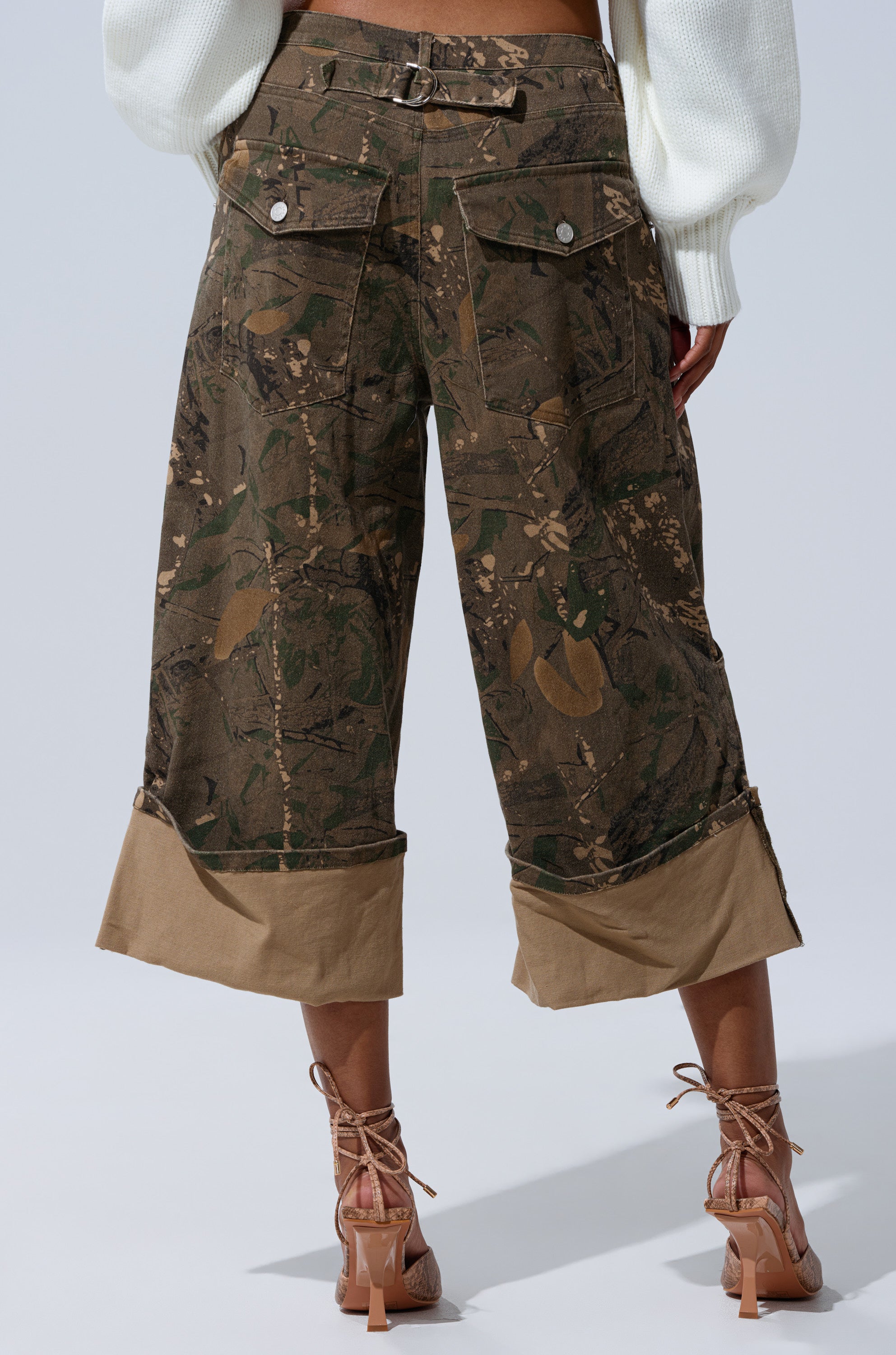 A person wearing DOWN AND DIRTY CAMO BERMUDA SHORTS with mid-rise fit, camouflage print, wide beige cuffs, large back pockets, and a belt detail, paired with tan high-heeled sandals and a white sweater. Photo taken from behind. 4