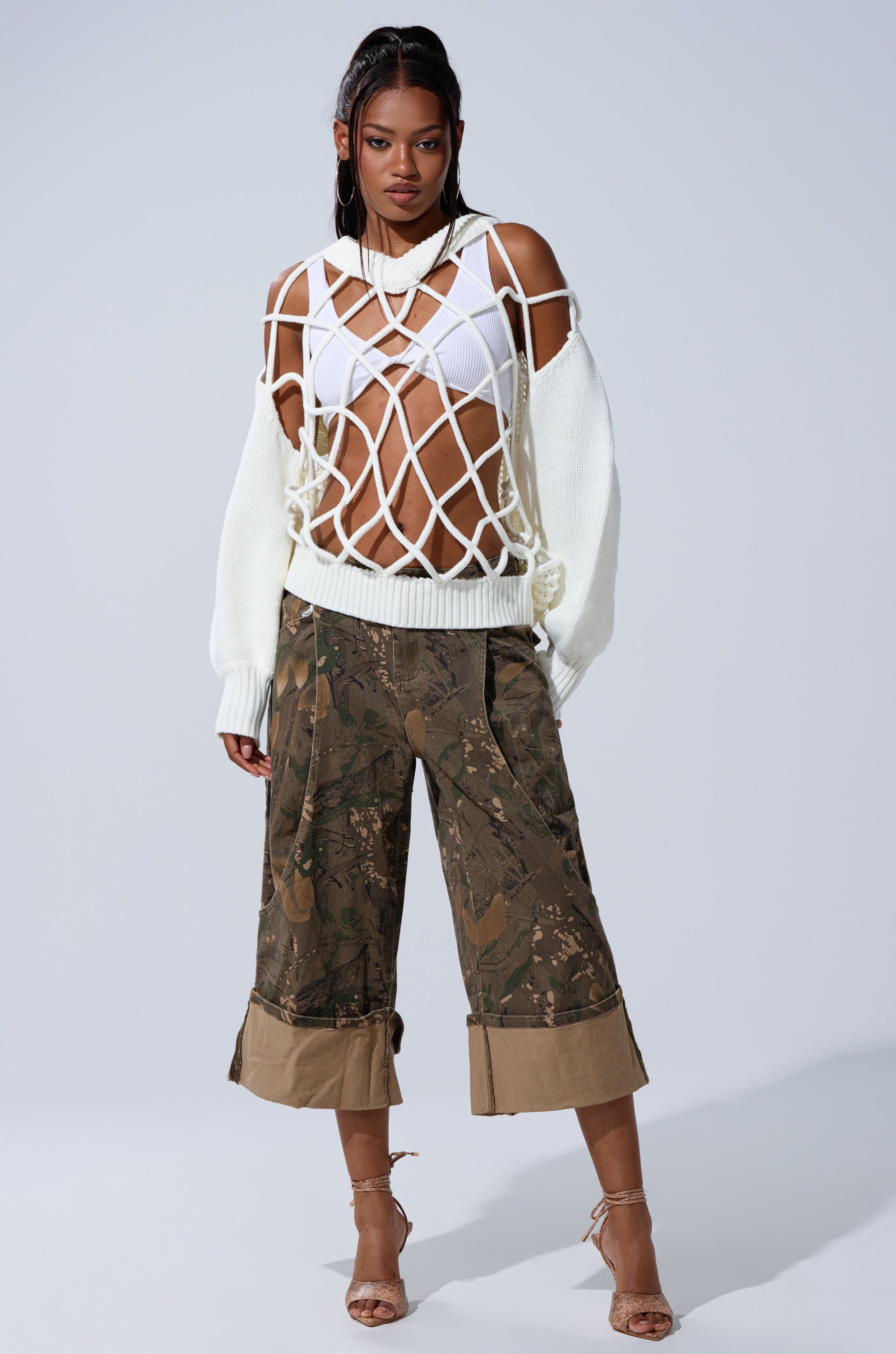 A woman poses confidently in the DOWN AND DIRTY CAMO BERMUDA SHORT paired with a white knit sweater, white bra, strappy heels, layered necklaces, and hoop earrings against a plain background. 5