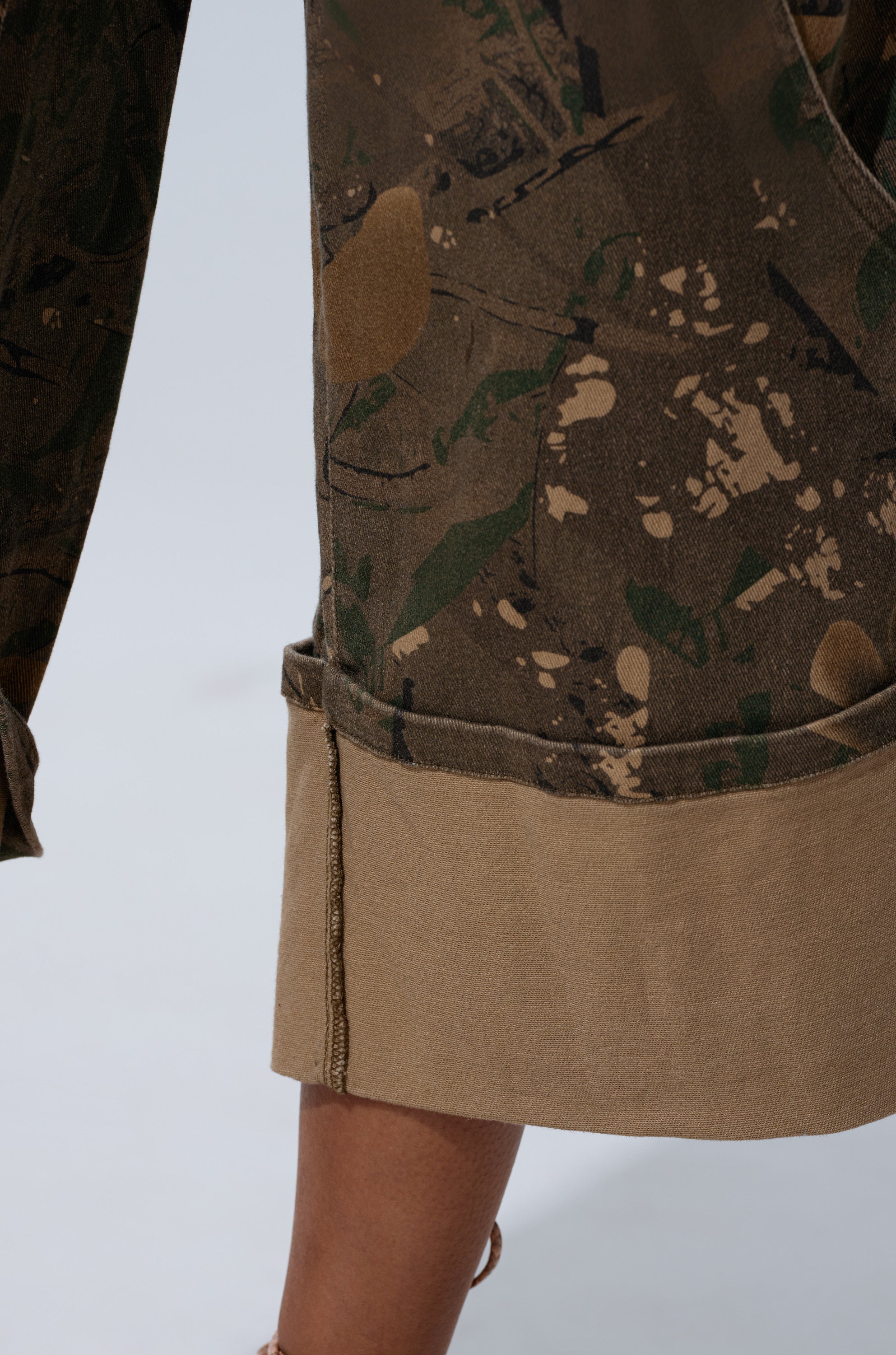 A close-up of a leg wearing the DOWN AND DIRTY CAMO BERMUDA SHORT, featuring a wide tan cuff at the hem and a comfy mid-rise fit. The plain background highlights its utilitarian style. 6