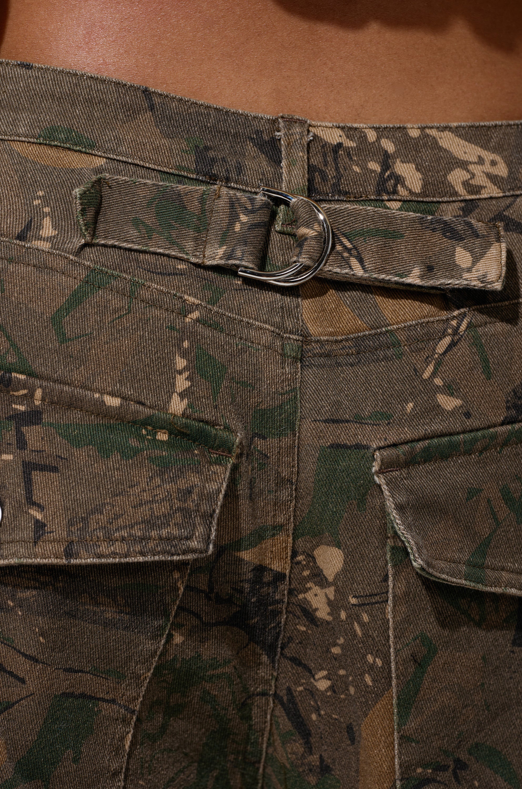 Close-up of the DOWN AND DIRTY CAMO BERMUDA SHORT shows back flap pockets and a silver D-ring belt detail below the waistband. The mid-rise fit and rugged camo texture combine green, brown, and tan tones for a utilitarian vibe. 7