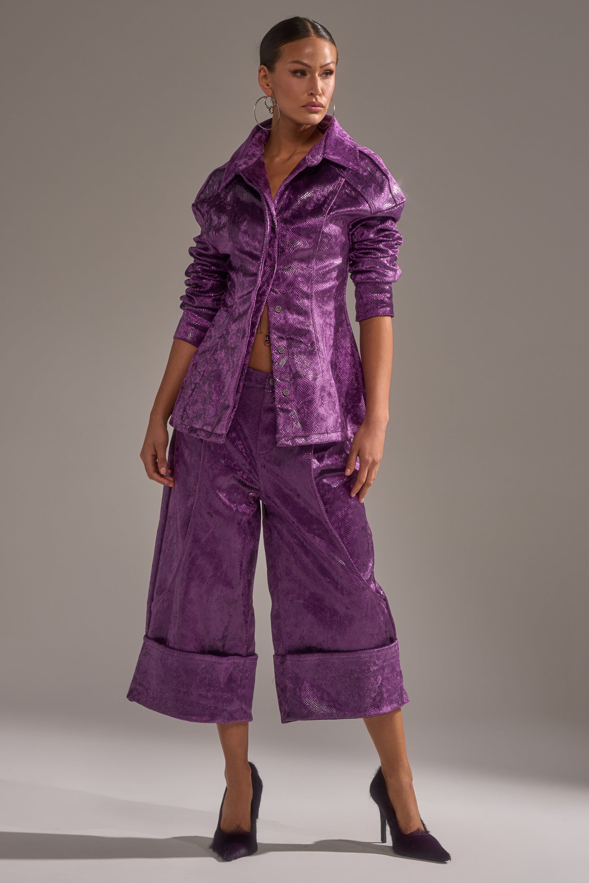A woman poses against a gray background wearing the CRAZY TOUCH VELVET TROUSER, a shiny purple velvet wide-leg culotte, paired with a fitted jacket and matching purple heels.