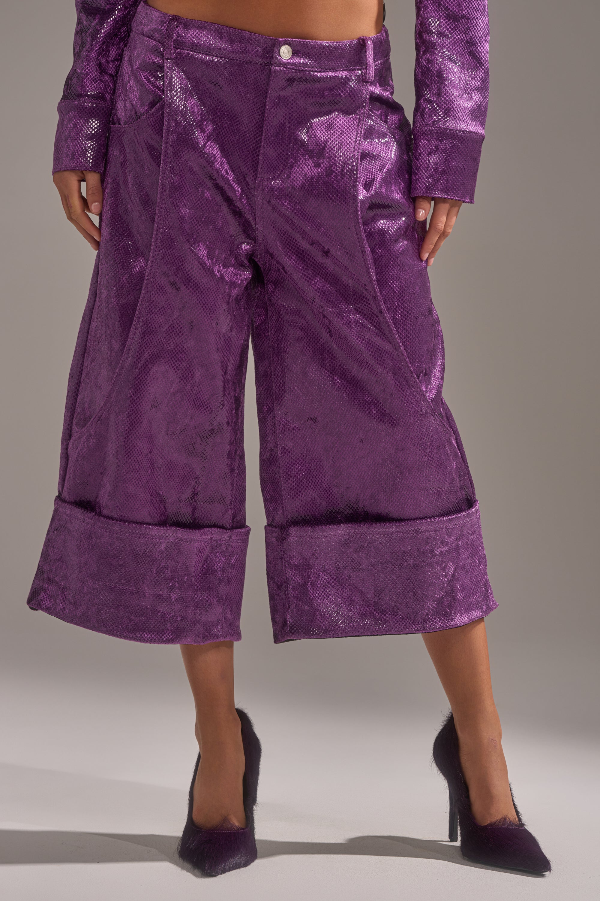A person wearing the CRAZY TOUCH VELVET TROUSER in shiny, textured purple with large cuffs and matching high heels stands against a neutral background.