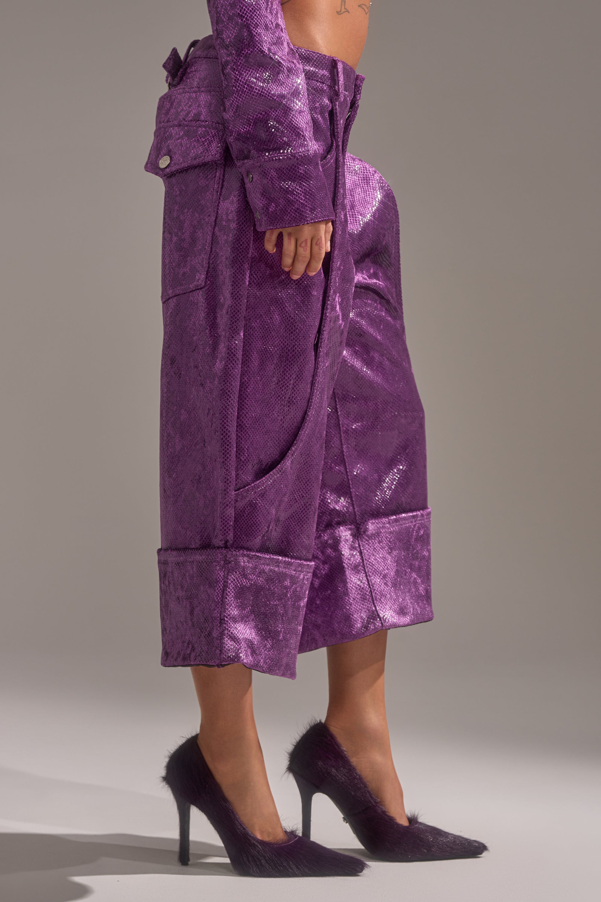 A person stands against a plain gray background wearing shiny purple CRAZY TOUCH VELVET TROUSER culottes with wide cuffs and matching pointed high heels covered in purple faux fur.