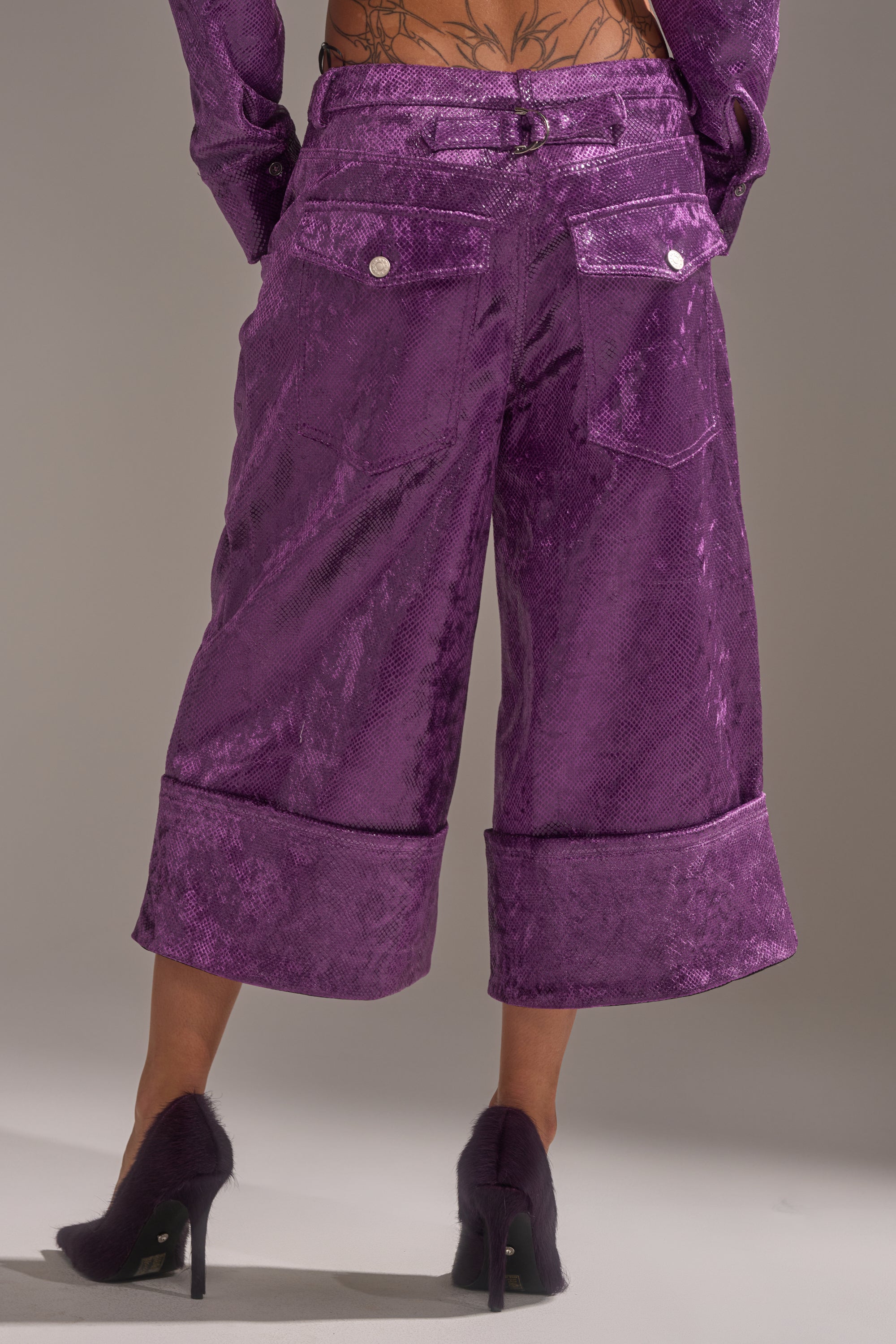 A person faces away, showcasing the CRAZY TOUCH VELVET TROUSER in shiny purple with large cuffs and matching high heels. The textured velvet fabric and their lower back tattoo are visible.