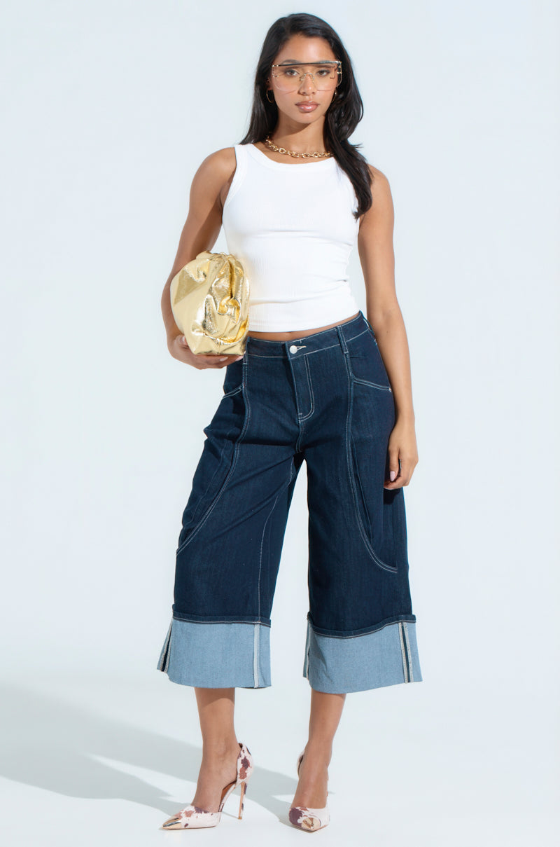 A woman stands against a light background in a sleeveless white top, COME WITH ME DENIM BERMUDA SHORT, and high heels. She holds a shiny gold purse, wears glasses, and lets her long hair down.