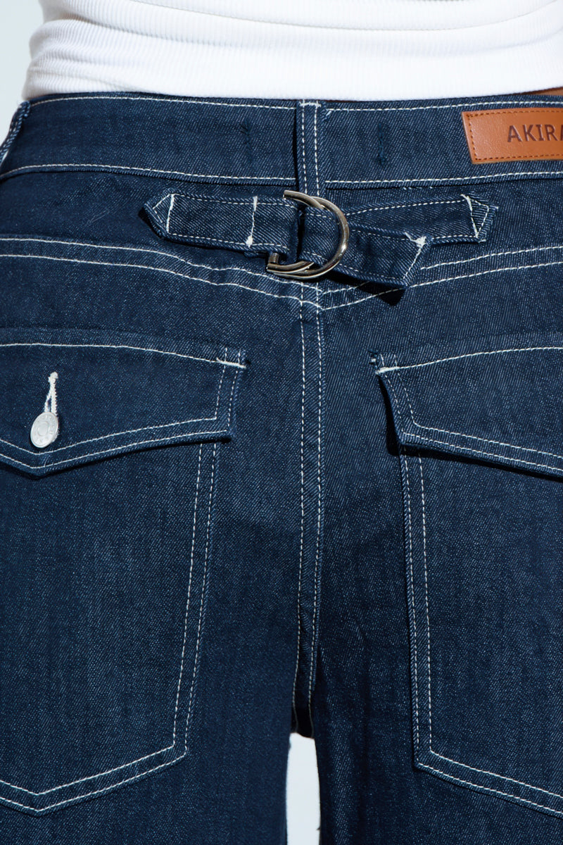 Close-up of the COME WITH ME DENIM BERMUDA SHORT in dark blue with large pockets featuring white-stitched buttoned flaps, a tan "AKIRA" leather patch, and curved panel construction. Made from a stretchy cotton blend, these shorts have belt loop details above the pockets.