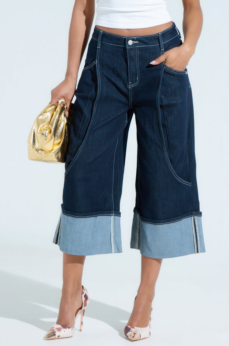Someone is dressed in COME WITH ME DENIM BERMUDA SHORT with a wide light blue hem, a white top, floral high heels, and carrying a shiny gold handbag for added glamour. The subtle stretch of the cotton blend ensures comfort against a light gray background.