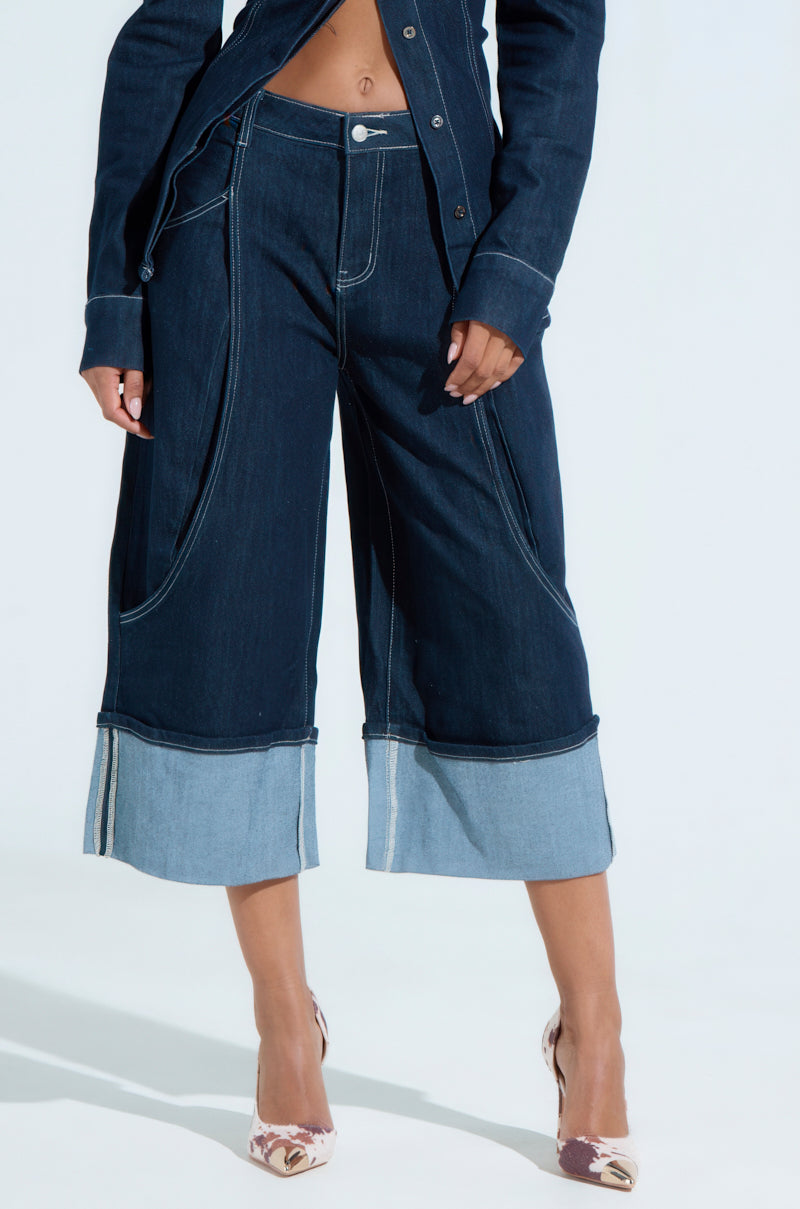 Someone wearing dark wide-legged denim pants with light-colored cuffs and made from a stretchy cotton blend, matched with a denim jacket, and styled with patterned high heels stands against a plain white backdrop.