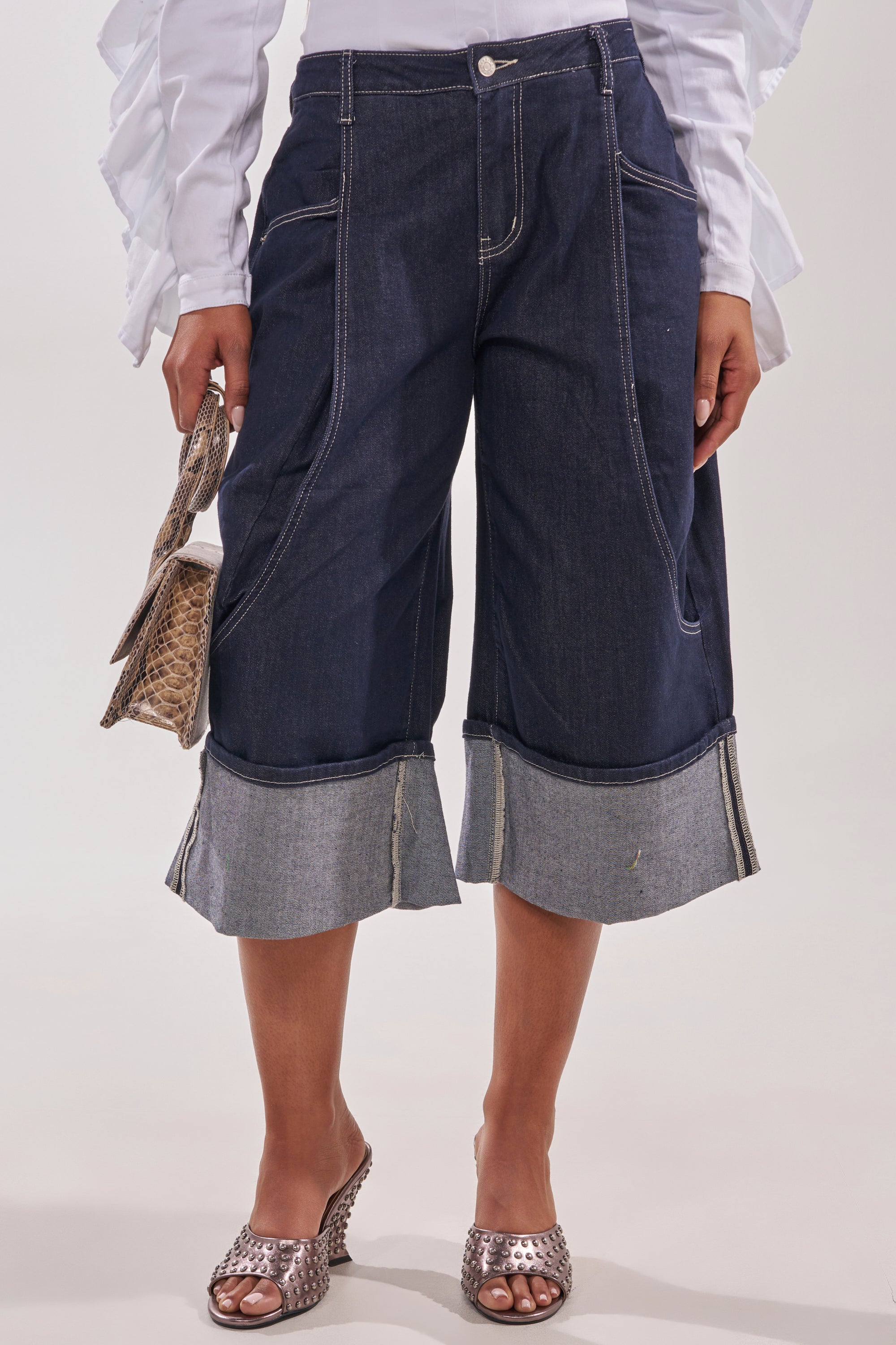 A person wearing the COME WITH ME DENIM BERMUDA SHORT styled with a white ruffled long-sleeved blouse, metallic open-toe heels, and holding a small textured handbag. 2