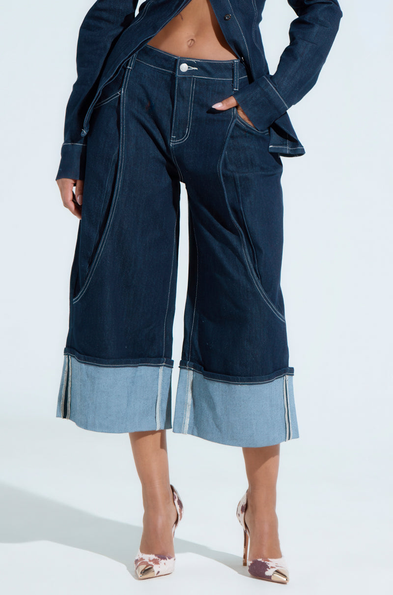 The person is sporting COME WITH ME DENIM BERMUDA SHORTS with a matching denim jacket, hands casually tucked in the pockets. They're paired with heels and crafted from a stretchy cotton blend for comfort, featuring a dark denim color with lighter denim at the bottom hem.