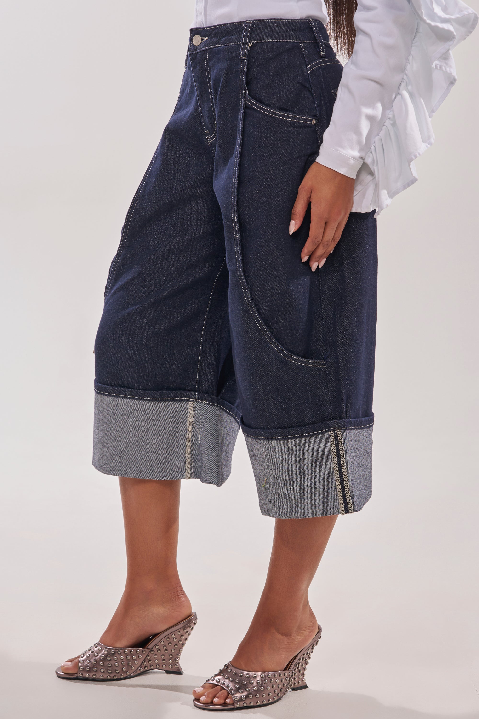 A person wears the COME WITH ME DENIM BERMUDA SHORT, featuring wide legs, dark denim, large front pockets, and light cuffs, styled with a white ruffled blouse and wedge sandals for a chic festival look. 3