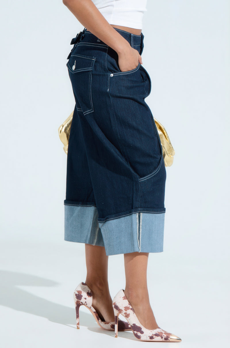 A person in a white top models the COME WITH ME DENIM BERMUDA SHORTS, standing sideways. One hand is in a pocket, the other holds a gold bag, and they wear white and brown high heels. The stretchy cotton blend offers comfort against a neutral background.