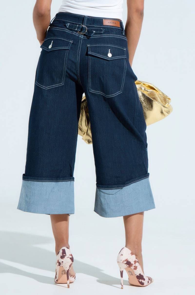 Rear view of a person wearing the COME WITH ME DENIM BERMUDA SHORT, crafted from a stretchy cotton blend with turned-up cuffs. They hold a gold bag and pair it with white high heels featuring brown patterns. The shorts have two back pockets and a leather patch on the waistband.