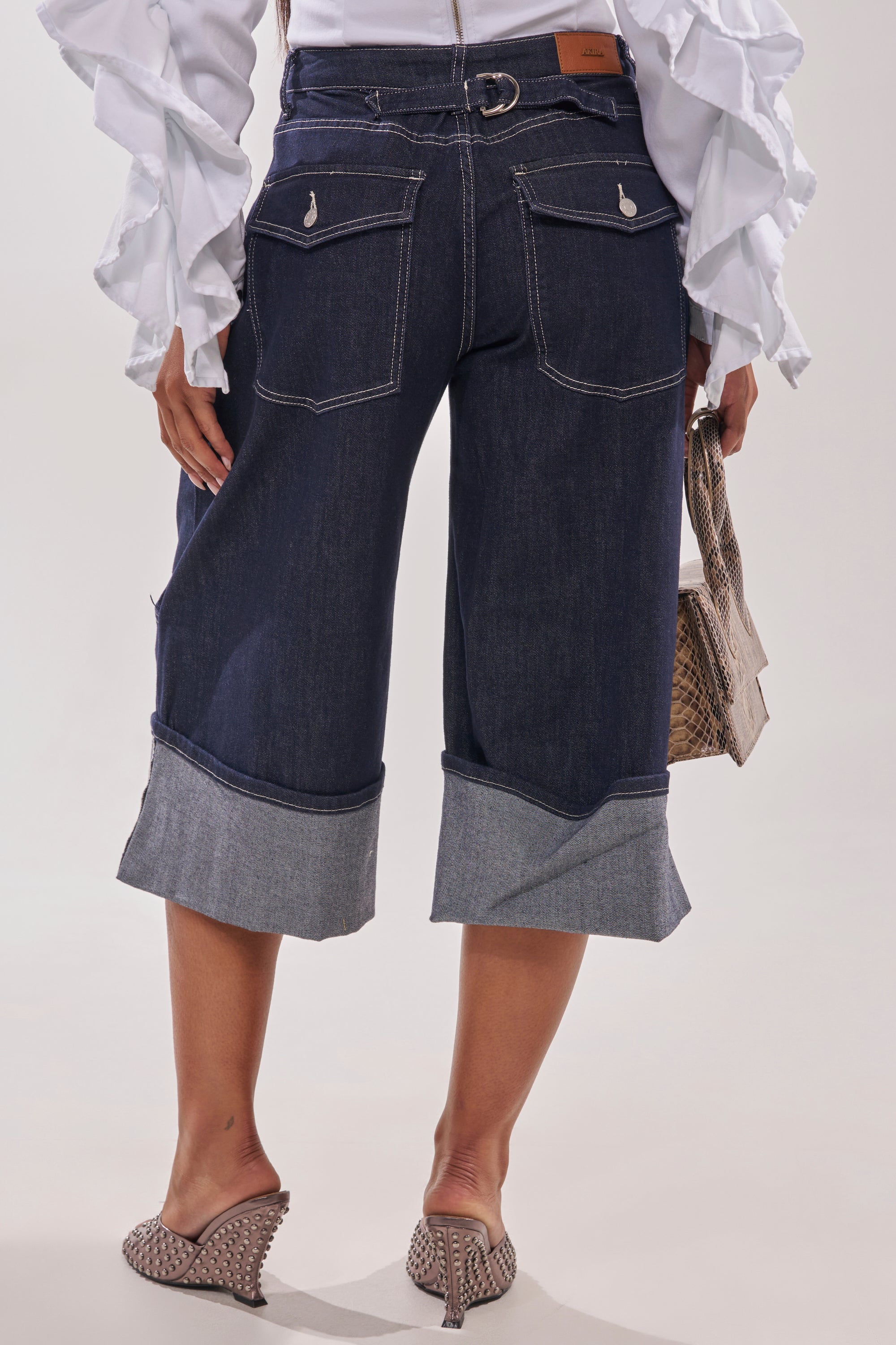 A person stands facing away, wearing the COME WITH ME DENIM BERMUDA SHORT, a white ruffled long-sleeve blouse, studded heeled mules, and holding a small textured handbag. 5